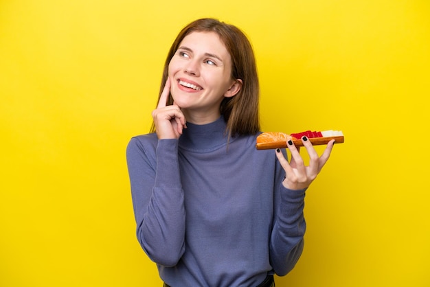 Young English woman holding sashimi isolated on yellow background thinking an idea while looking up