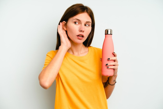 Young English woman holding a thermo isolated on blue background trying to listening a gossip.