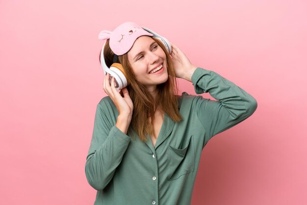 Young English woman in pajamas isolated on pink background in pajamas and holding a pillow and listening music