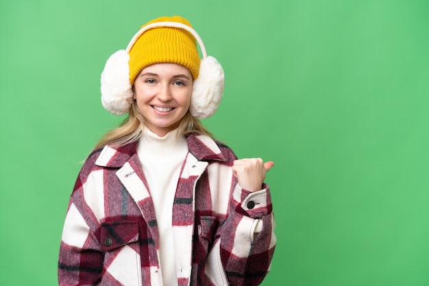 Young English woman wearing winter muffs over isolated background pointing to the side to present a product