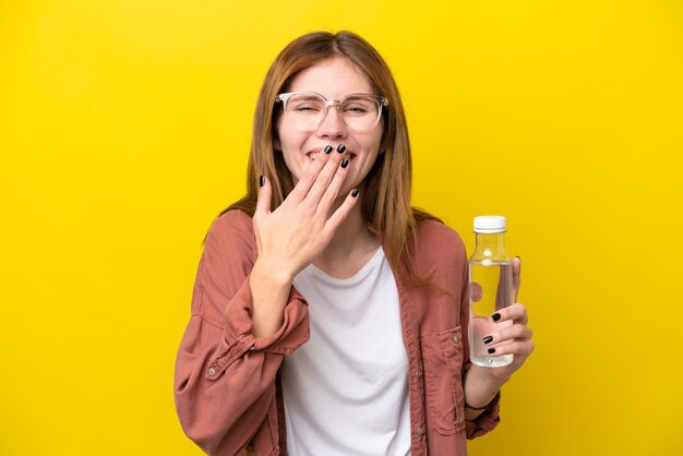 Young English woman with a bottle of water isolated on yellow background happy and smiling covering mouth with hand