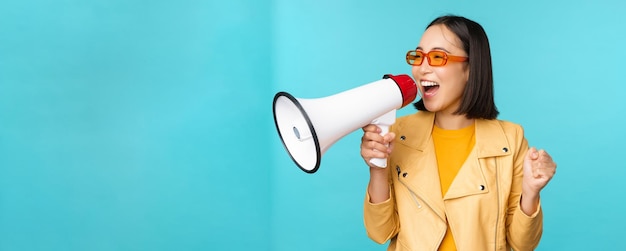 Young enthusiastic korean girl in trendy outfit shouting in megaphone making announcement advertising screaming in speakerphone standing over blue background