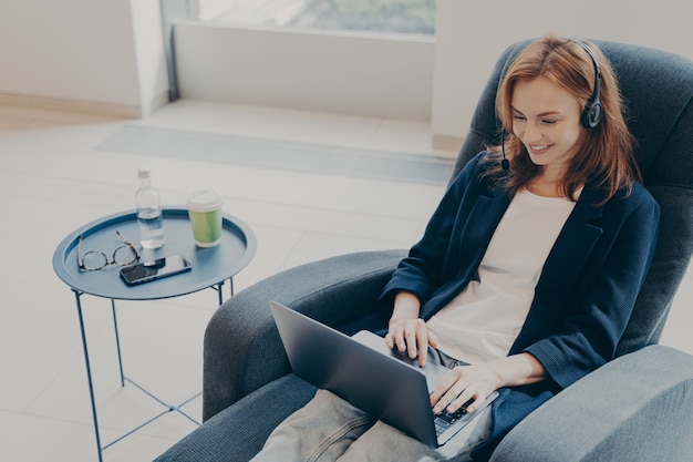 Young entrepreneur lady wearing headset using laptop while sitting in armchair at home