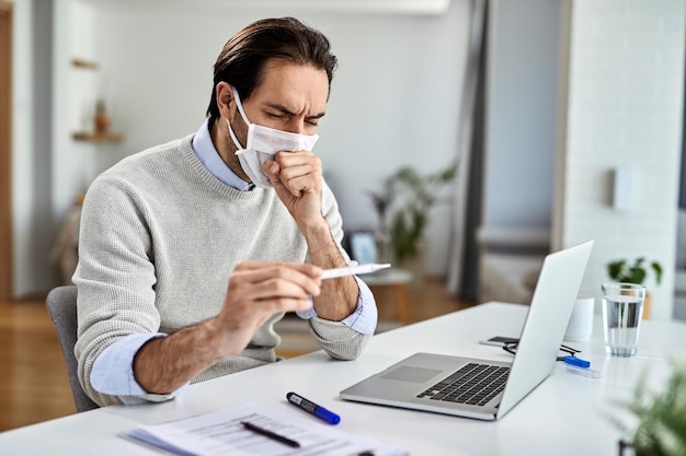 Young entrepreneur wearing protective face mask and coughing while measuring temperature at home