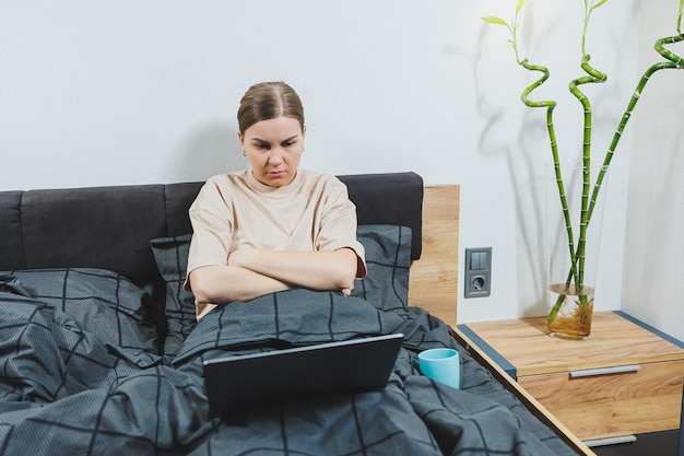 Young European beautiful woman sitting on bed with coffee working with computer at home remote work Woman happy and smiling in bed at home