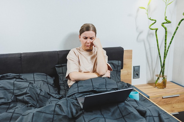 Young European beautiful woman sitting on bed with coffee working with computer at home remote work Woman happy and smiling in bed at home