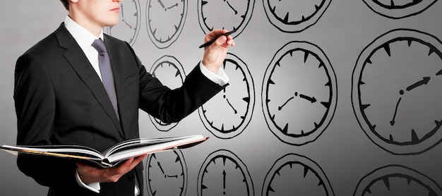 Young european businessman with open book standing on concrete wall background with creative hand drawn clock sketch Time management deadline and career concept