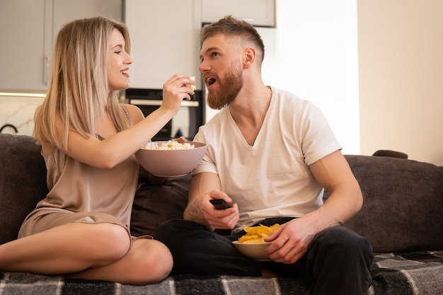 Young european couple sitting on sofa and watching TV or movie. Blonde girl feeding popcorn of her boyfriend. Leisure and rest at home. Concept of enjoying time together. Interior of studio apartment