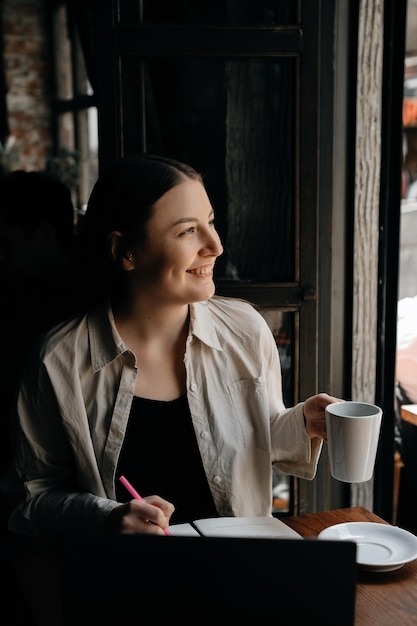 A young European freelancer girl with long dark hair drinks coffee and remotely works in a cafe