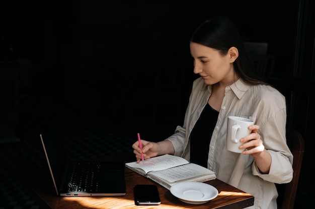 A young European freelancer girl with long dark hair remotely works and studying in a cafe