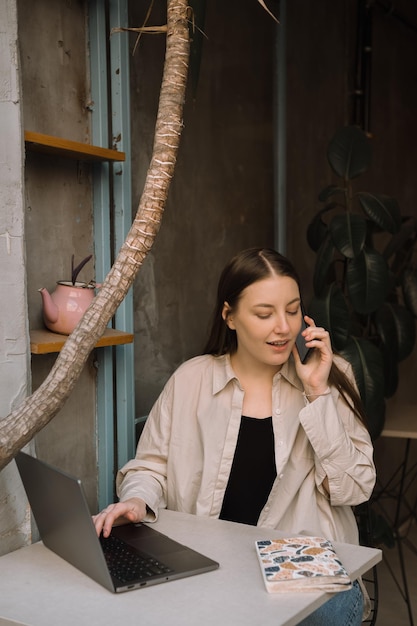 A young European freelancer girl with long dark hair in a shirt and jeans remotely works in a cafe