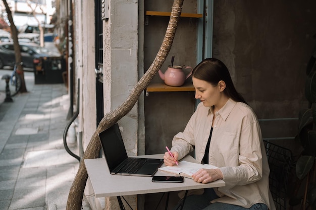 A young European freelancer girl with long dark hair in a shirt and jeans remotely works in a cafe