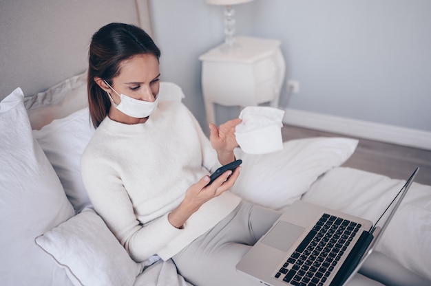 Young european woman in face mask in bedroom with laptop during coronavirus isolation