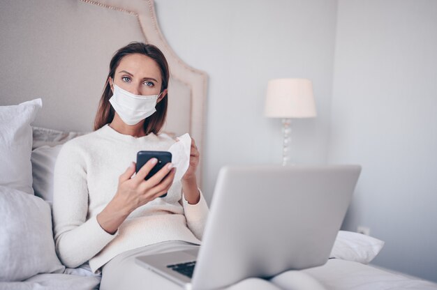 Young european woman in face mask in bedroom with laptop during coronavirus isolation