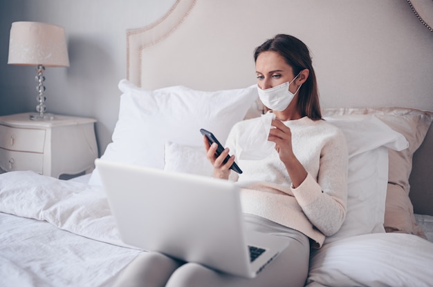 Young european woman in face mask in bedroom with laptop during coronavirus isolation