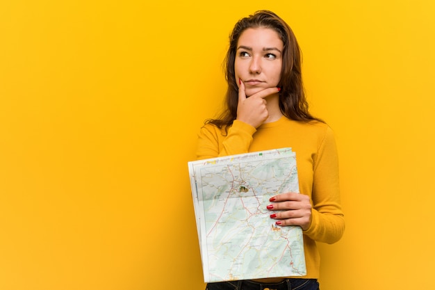 Young european woman holding a map looking sideways with doubtful and skeptical expression.