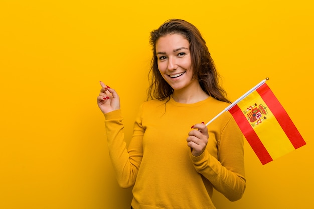 Young european woman holding a spanish flag smiling cheerfully pointing with forefinger away.