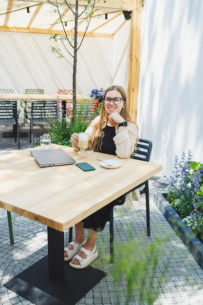 Young European woman sitting at outdoor cafe table with laptop and cup of coffee smiling woman in glasses enjoying telecommuting in cafe or studying online