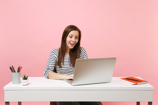 Young excited amazed woman in casual clothes working on project with laptop while sit at office isolated on pastel pink background. Achievement business career concept. Copy space for advertisement.