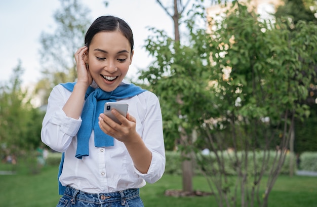 Young excited Asian woman holding smartphone, using mobile app for online shopping