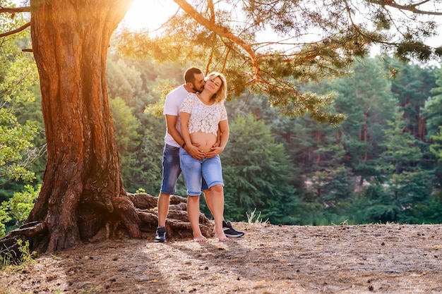 Young expecting couple holding hands on belly under the tree at sunset