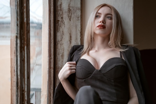 Young fairhaired woman wears black outfit bustier with sweetheart deep neckline sitting on the windowsill Sad girl