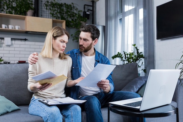 Young family husband and wife reviewing their bills loan and mortgage agreements sitting on the couch at home working with documents on a laptop embarrassed upset