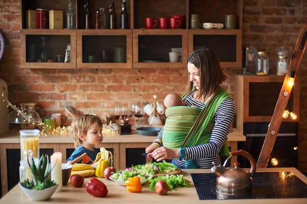 Young family mother with two children preschool boy and baby in sling cooking together in a sunny kitchen