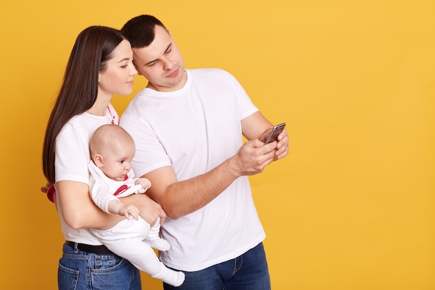 Young family smiling while looking together at smart phone screen, husband showing something interesting to his wife, posing with newborn daughter against yellow wall