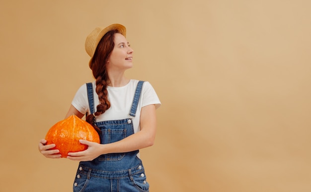 Young farmer woman in overalls holds a ripe orange pumpkin on a yellow background