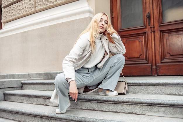 Young fashion blonde woman in a trench coat sits on stairs in the city