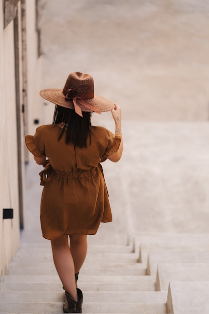 Young fashion woman dressed in a summer dress with a wicker wide hat walks and poses in the old city.