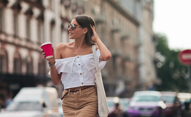 Young fashion woman in trendy summer clothes holding coffee cup while walking around the city