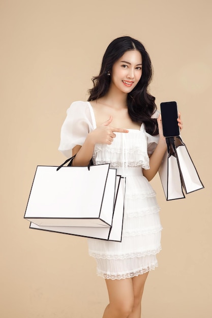 Young fashionable asian woman showing blank screen mobile phone holding shopping bags isolated on beige background and copy space Shopping online payment on smartphone