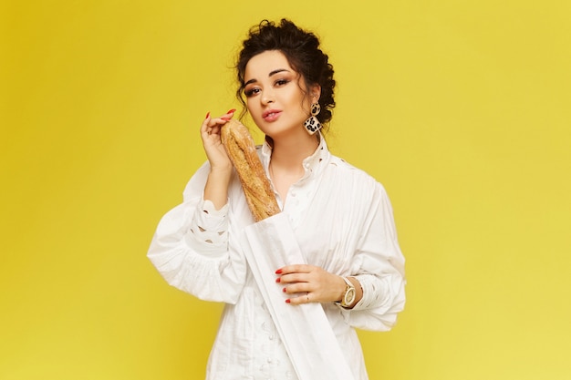 Young fashionable lady wearing white summer dress posing with crispy baguette over the yellow backgr...