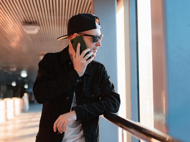 Young fashionable modern guy a man in a black cap and sunglasses with a smartphone talks on the phone on a city urban background in a tunnel in the sunset rays of the sun