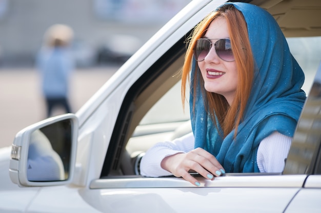 Young fashionable smiling woman driver looking out the window behind car steering wheel.