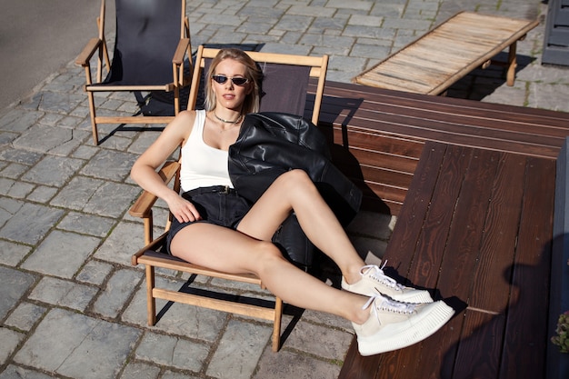 young fashionable woman relaxing on chaise longue wearing stylish sunglasses shorts sneakers