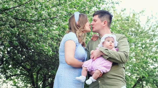 Young father in green shirt holding baby girl kisses mother wearing summer dress in city park against blossoming fruit trees with green leaves