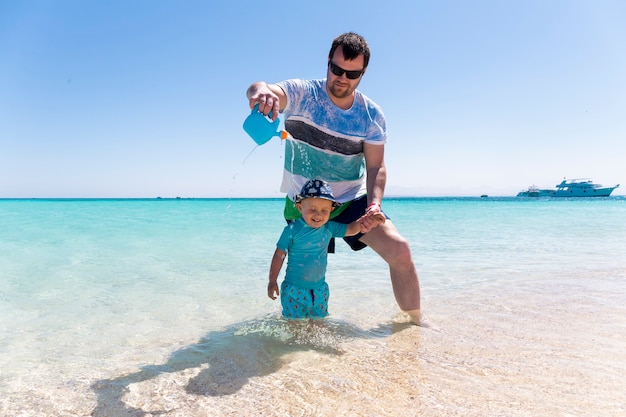 A young father is playing on the beach with his little baby son and waters him from the watering can. Little boy is laughing at the seaside.