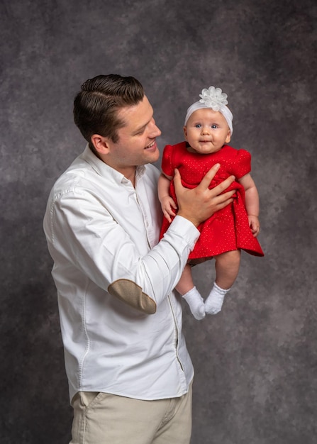 A young father in a light shirt gently holds his little daughter dressed in a red dress in his arms Studio shooting Father's love for newborn baby