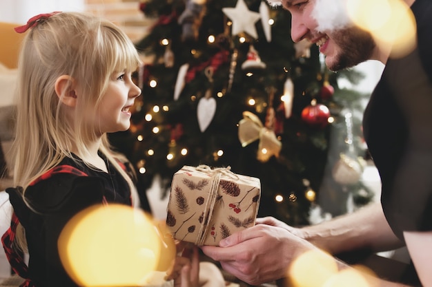 Young father in red Santa hat giving Christmas gift to his smiling daughter near decorated Christmas tree. Girl dressed in festive red-black Christmas outfit. They smiling, happy because of New Year