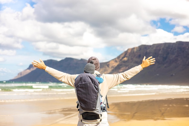 Young father rising hands to the sky while enjoying pure nature carrying his infant baby boy son in backpack on windy sandy beach of Famara Lanzarote island Spain Family travel concept