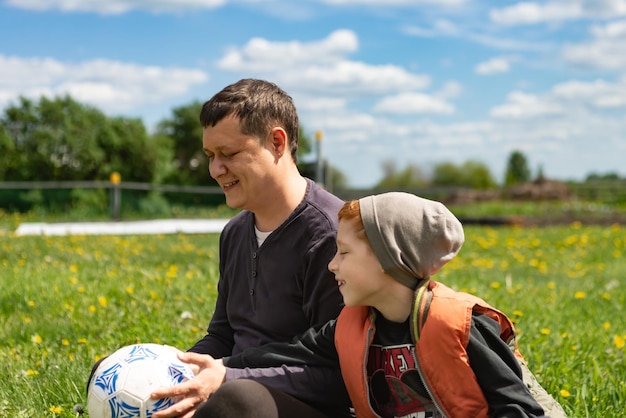 Young father and son sit on the grass in summer after playing football time together lifestyle portrait