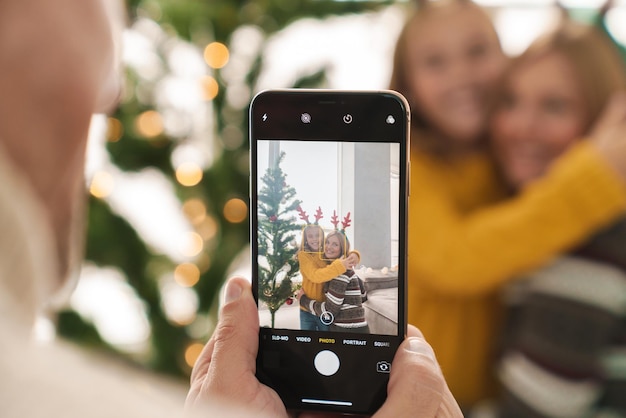 Young father taking a picture with mobile phone of his wife and daughter hugging at the christmas tree, celebrating holiday