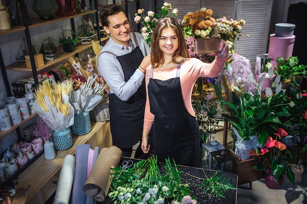 Young femal and male florists stand in room full of flowers and plants. She hold hone. They take selfie. People smile and pose.