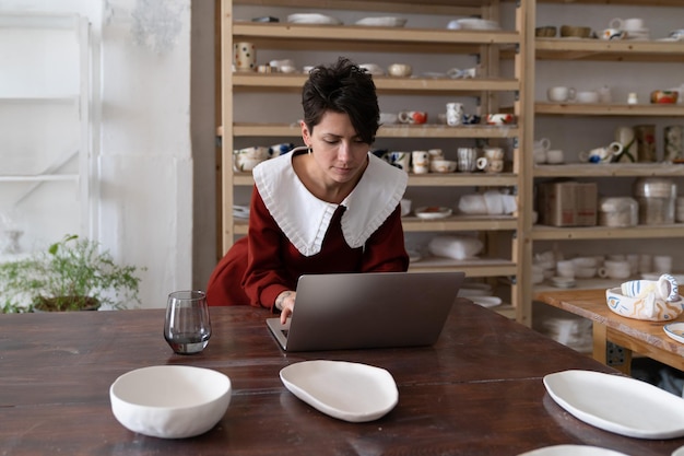 Young female artisan working on laptop in pottery studio making money with ceramics