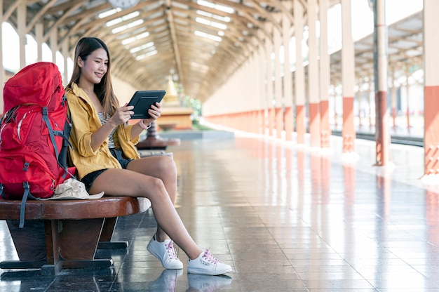 Young female backpacker using tablet searching destination to go while sitting at railway station