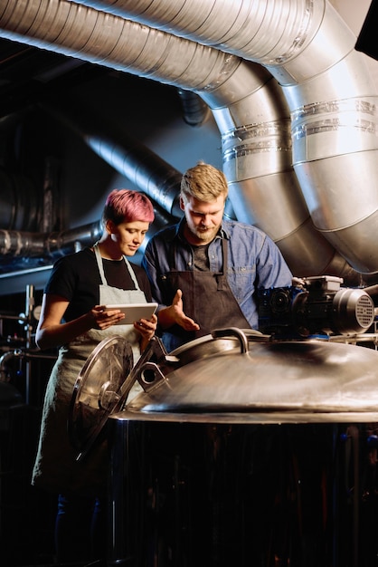 Young female brewer with digital tablet standing by her male colleague while both using equipment for beer preparation inside large workshop