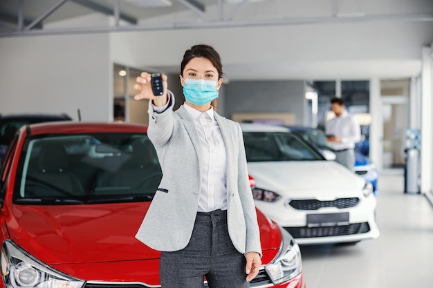 Young female car seller with face mask standing in car salon and showing car keys